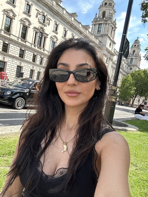 The image features a woman wearing sunglasses, posing for a photo with her arm extended outwards. She has long hair that falls over her shoulders. The background of the picture includes a street scene with cars parked along the side, as well as some buildings in the distance. There is also an element of a car and a person sitting on a bench near one of the buildings. The image seems to be related to a post about a millionaire crashing out on tl or a bandit & Pingu having an affair, with the woman's pose suggesting that she might have been involved in such events.