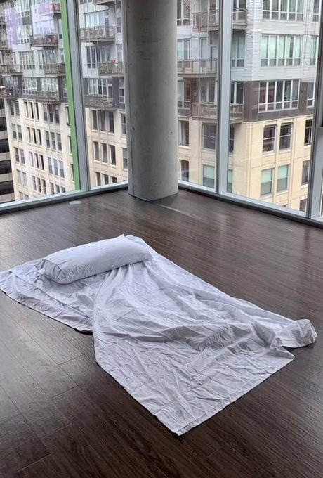 A thin mattress sheet and pillow lie on the floor of a high-rise apartment with floor-to-ceiling windows overlooking a city skyline, illustrating the poster's financial misfortune from buying SOL at $250.