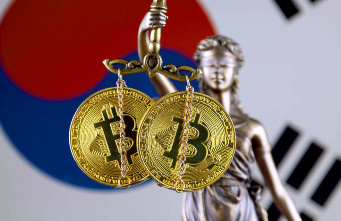 Gold Bitcoin coins and chains are draped over a scale of justice statue with the South Korean flag in the background, representing cryptocurrency regulation.