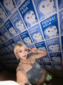 A woman with blonde hair poses for a selfie in a denim top against a wall covered in 'PENGU ASIA' and 'JONI THE' penguin posters.