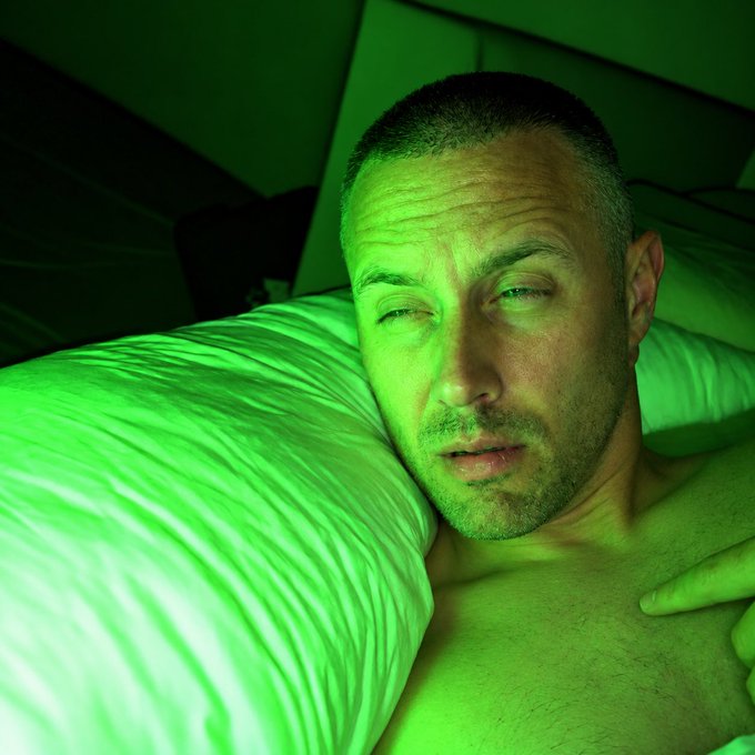 A close-up selfie of a man with short hair resting his head on a pillow, illuminated by strong green lighting in a dark room.