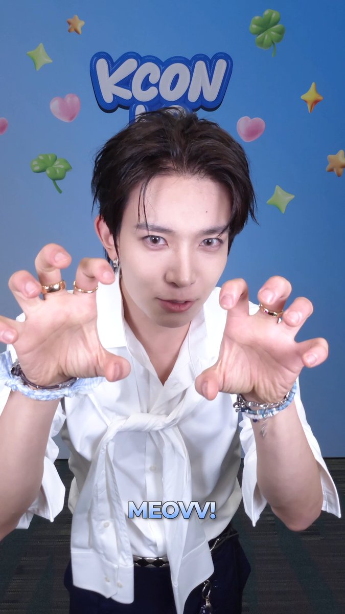 A young man with dark hair makes playful "clawing" gestures toward the camera, wearing a white knotted shirt, silver rings, and bracelets against a blue "KCON" backdrop decorated with stars, hearts, and clovers, with the caption "MEOVV!" at the bottom—capturing a fun roll-call moment at KCON LA 2026.