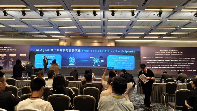 A conference hall where attendees sit facing a stage presentation titled 'AI Agent: 从工具到参与者的演化 (From Tools to Active Participants)', featuring slides about AI agents and LLM architecture.