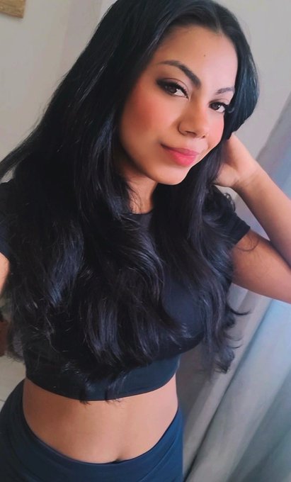 A woman with long, wavy black hair poses in a black crop top, smiling subtly for the camera in a relaxed selfie. The casual, confident vibe perfectly matches her "Happy Friday Friends" post, capturing a fun and carefree start to the weekend.