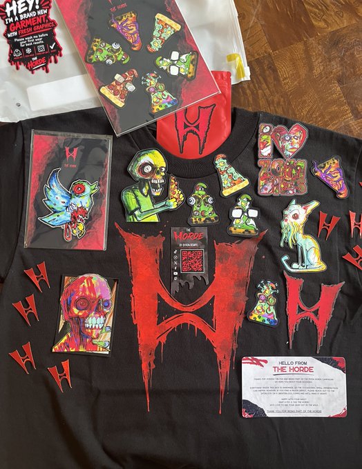 A black t-shirt displayed with horror-themed 'THE HORDE' merchandise including colorful zombie/skull enamel pins, a trading card, and stickers arranged on top, along with promotional tags.