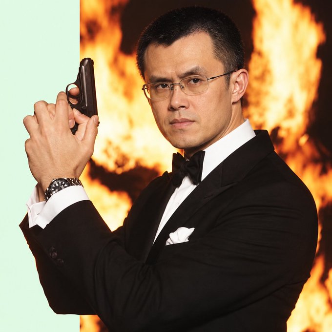 A man wearing a tuxedo with a bow tie and glasses, pointing a pistol with a serious expression against a backdrop of flames on one side and a light green background on the other.