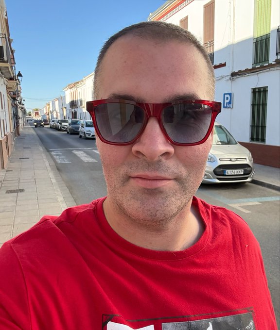 A selfie of a man with short hair wearing red sunglasses and a red t-shirt, standing on a sunny street with white buildings, parked cars, and a 'P' parking sign visible in the background.