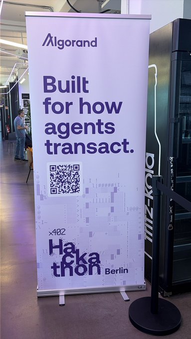 The image features a large white banner with black text that reads "Built for how agents transact. Hackathon Berlin". The banner is displayed prominently, possibly at an event or gathering related to the hackathon. There are two people visible in the background of the image, one standing near the left side and another person closer to the center.