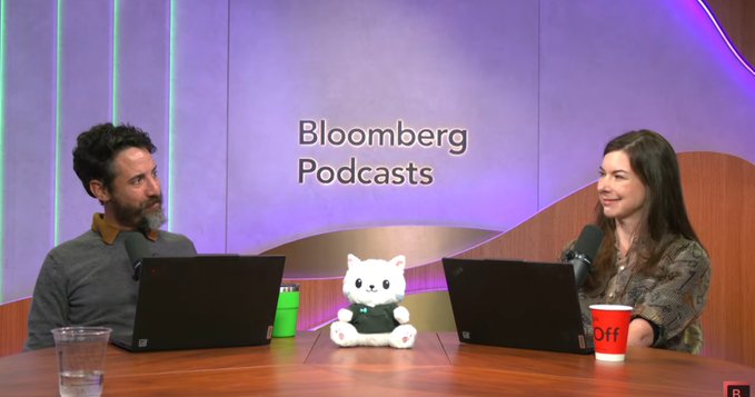 The image shows two podcast hosts sitting at a desk with a small white cat plush toy (Hypurr) between them, with "Bloomberg Podcasts" displayed on the backdrop, visually representing the Odd Lots discussion about Hyperliquid's mascot.