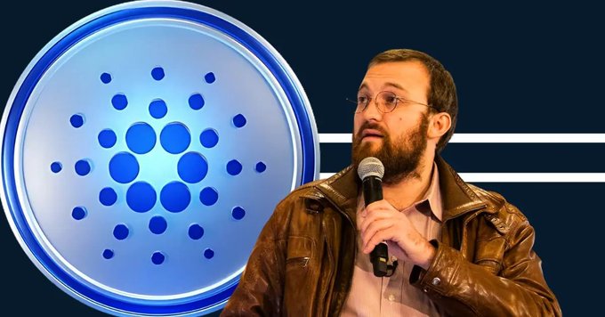 A bearded man wearing glasses and a brown leather jacket speaks into a microphone while standing next to a large blue circular logo resembling the Cardano cryptocurrency emblem.