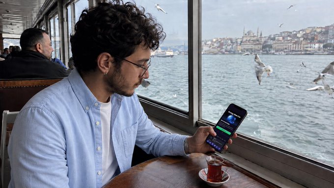 A young man with glasses and a beard sits by a window looking at his smartphone, with a view of seagulls flying over water and a city skyline in the background, and a glass of tea on the table.
