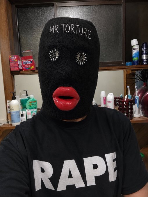 The image features a person wearing a black mask with red lips, which has the words "Mr Torture" written on it. The person appears to be posing for a photo or selfie, as indicated by their facial expression. The background of the image includes various bottles and other items typically found in a bathroom setting.