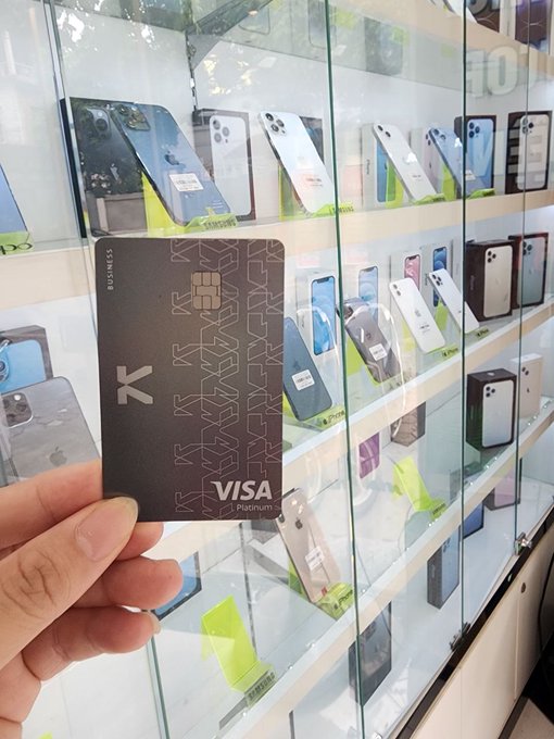 The image shows a person holding up a Visa card, which has a black background and white text that reads "Visa". The person's hand is visible above the card as they hold it up against a backdrop of cell phones displayed on shelves or in cases behind them. This suggests that the person might be interested in purchasing one of these devices using their Visa card.