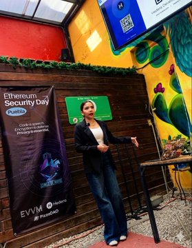 The image features a woman standing in front of a wooden wall, giving a presentation or speech. She appears to be wearing a black blazer and blue jeans. The background includes a TV screen displaying a QRCode, possibly related to the event she is speaking at. There are also two signs on the wall that read "EVM" and "Hedera", which could potentially be related to the community or organization she represents.
