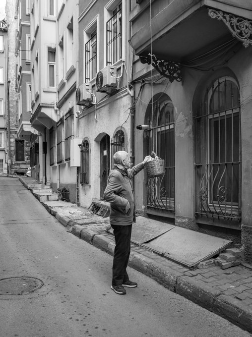 Black and white photo of an elderly man on a narrow street lowering a basket from a window using a rope, likely receiving groceries from above.