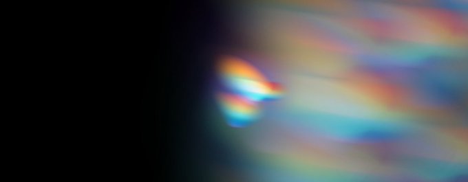 A colorful, prismatic light flare with a bright central beam and iridescent spectrums of pink, blue, and orange streaks against a dark background, visually evoking the ethereal, glowing aesthetic of the AuraSea NFT launchpad.