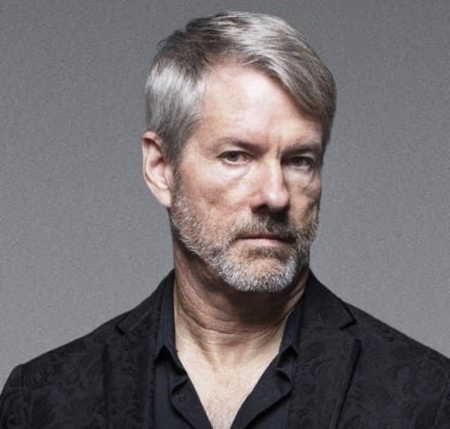 A professional headshot portrait of a gray-haired, bearded man wearing a black shirt and dark patterned jacket against a gray background.