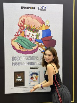 A woman in a black and red dress posing next to a KAKAWON 2026 promotional poster featuring the 'PUPPY/PENGLIN'S RACE' mascot design and branding from PT L7F and other sponsors.