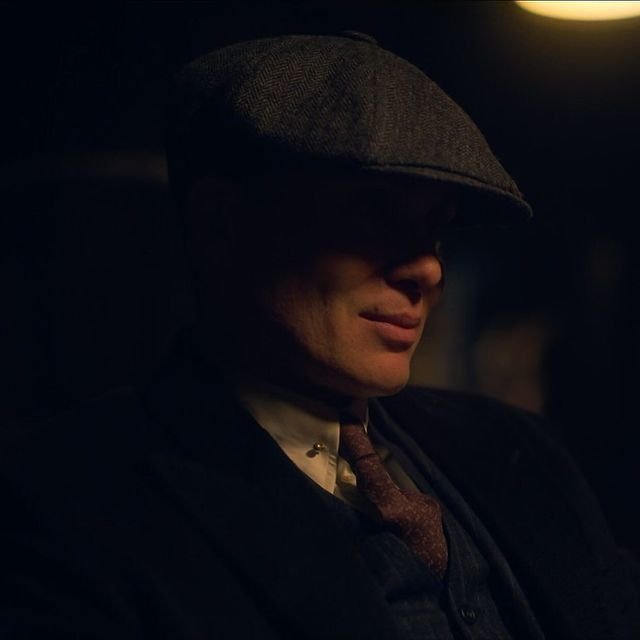 A moody, low-lit cinematic portrait of a man wearing a flat cap and suit with a tie, reminiscent of the Peaky Blinders series style.