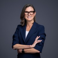 Cathie Wood, CEO of ARK Invest, stands confidently with her arms crossed in a professional navy blazer. Her composed, authoritative presence visually reinforces the post's message about her bold and consistent recommendation to invest in Bitcoin monthly.