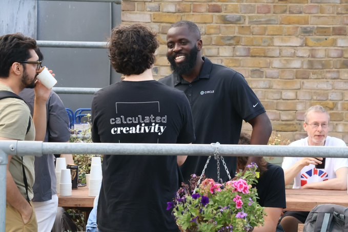 A smiling Black man wearing a black 'Circle' branded polo shirt converses with people at an outdoor gathering, while a man in a 'calculated creativity' shirt faces him.