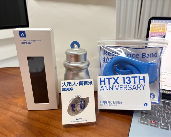 A collection of HTX 13th Anniversary merchandise is displayed on a desk, including a sports cooling towel, a water bottle, a resistance band, and a Trade Crypto Only On HTX card featuring a coin design with '火币人·真有米' text.