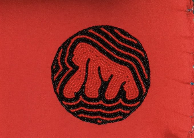 A circular beadwork patch on red fabric depicting a stylized bear or similar animal with black beaded outlines and red dotted fill.