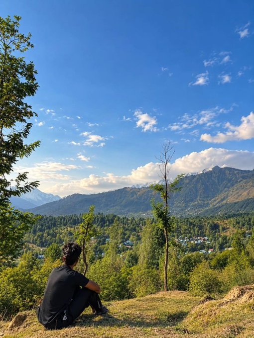 A person in dark clothing sits on a grassy hilltop overlooking a vast green mountain valley with a village below and a bright blue sky with scattered clouds.
