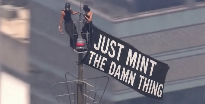 Two people on top of a tower or antenna holding a banner that reads 'JUST MINT THE DAMN THING'.