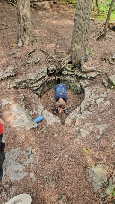 A child in a blue 'MIAMI' shirt climbing down into a rocky crevice in a forested area, with another person's leg visible at the edge.