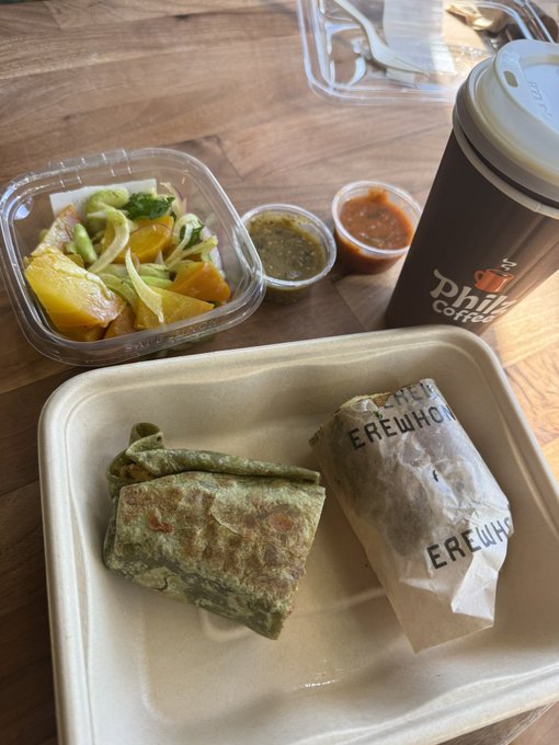 A breakfast spread featuring green spinach wraps—one sliced open and one wrapped in "EREWHON" branded paper—paired with a fresh fruit cup, salsa, and a Philz Coffee cup, visually echoing the post's "bull market breakfast" theme where green food mirrors bullish green market candles.