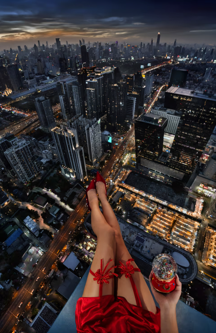 A woman in a red dress and high heels dangles her legs from the edge of a rooftop high above a sprawling city at dusk, holding a glittery cup while overlooking a vast urban skyline. The dramatic "on top of the world" perspective visually echoes the post's reference to the iconic Empire State Building setting, fitting the listing of a 1/1 piece originally purchased in 2023.