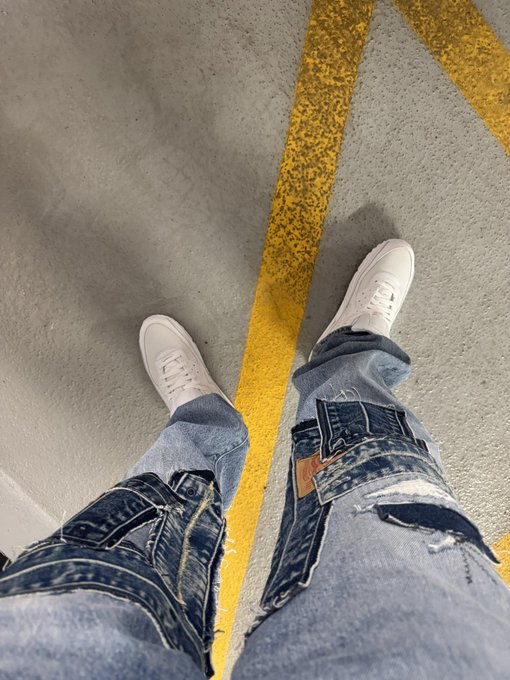 A first-person view shows someone wearing distressed denim jeans and white sneakers standing on a gray concrete floor marked with yellow lines. The "GM" (good morning) caption paired with the casual, freshly-outfitted look makes for a relaxed Wednesday greeting — looking sharp and ready for the day ahead.