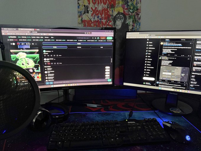 A dimly lit computer desk setup featuring dual monitors, a Dell keyboard, and a microphone, displaying what appears to be social media feeds on the screens.