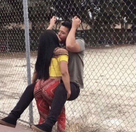 A young couple embraces and kisses against a chain-link fence in an outdoor setting, with the woman wearing a yellow top and red patterned leggings while the man in an olive shirt holds onto the fence. The post caption "TMA wallet watchers" humorously suggests the couple is so engrossed in each other that they're unaware of being watched, or that observers are noting (and possibly judging) their public display of affection.