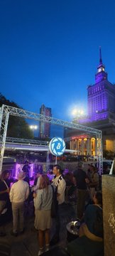 An evening outdoor event scene with people gathered near an illuminated structure, with a tall building lit in purple visible in the background.