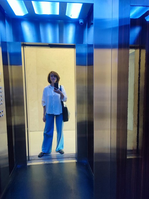 In this image, there is a woman standing inside an elevator with blue lighting. She is wearing a white shirt and blue pants. In her hand, she is holding a black purse. The background of the image features several windows that provide a view outside. On the walls, there are signs hanging on them.