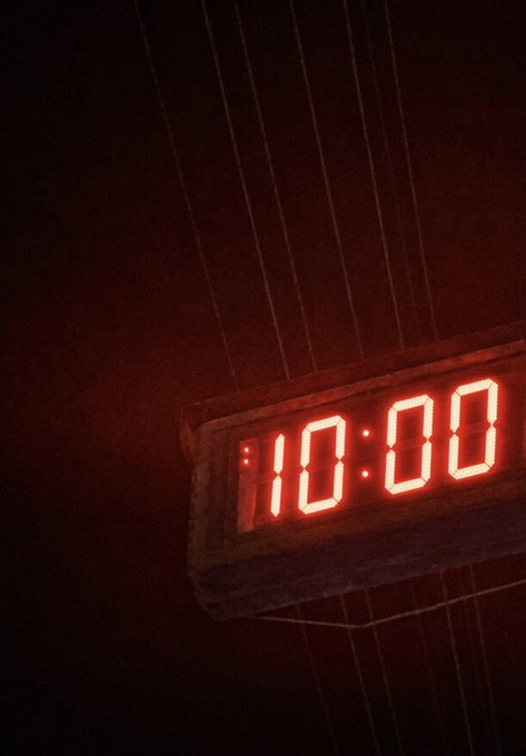 A glowing red digital clock displays '10:00' in a dark setting, with vertical cable-like elements visible in the background.
