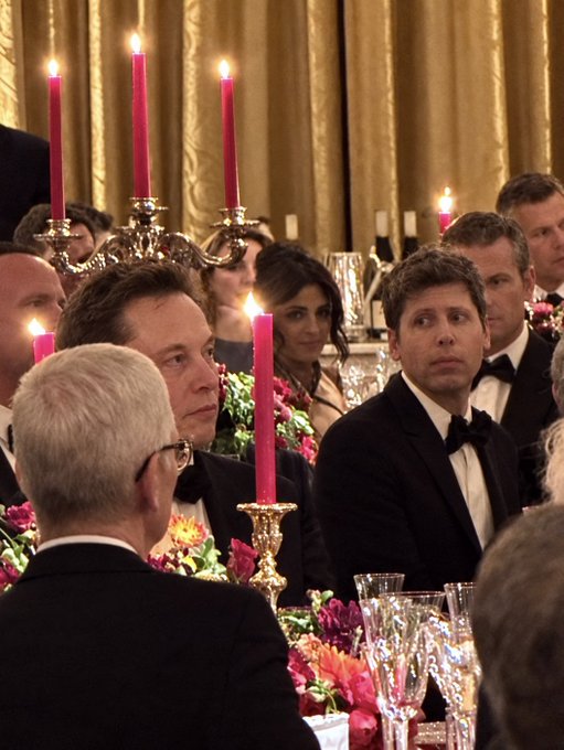 Elon Musk and Sam Altman sit side by side in black tuxedos at a formal candlelit dinner event, with tall pink candles and floral arrangements in the foreground.