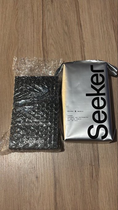 Two products from the brand 'Seeker' by Solana Mobile are displayed on a wooden floor: a black phone case in bubble wrap on the left and a silver/sealed package on the right with text 'SOLANA MOBILE, SEEKER, DESIGNED AND DISTRIBUTED BY SOLANA MOBILE, SEE, THE WORLD.'