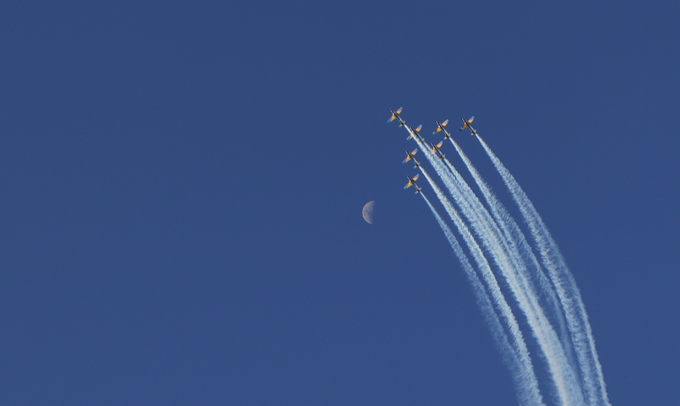 Seven jet aircraft fly in tight formation across a deep blue sky, leaving long white contrails behind them, with a small crescent moon visible in the distance.