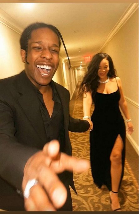 A man in a dark suit laughs joyfully in the foreground, extending his hand, while a woman in a black, high-slit dress smiles in the background hallway.