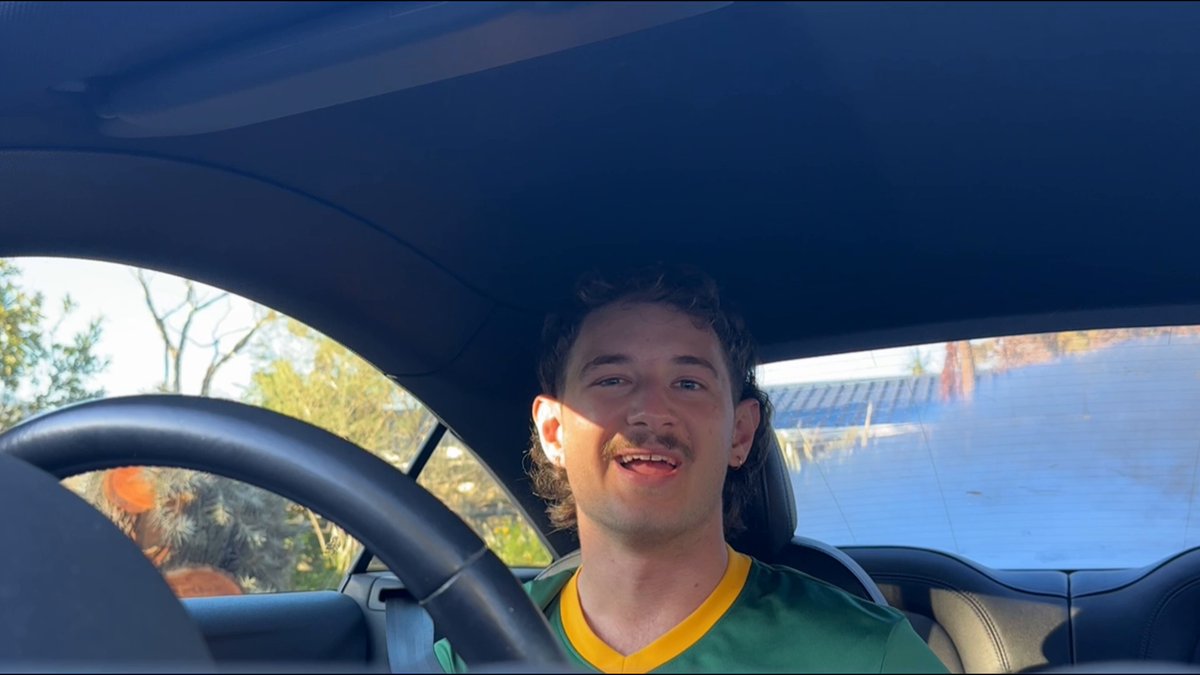 The image shows a man sitting inside a car, wearing a green and yellow shirt with his eyes closed as if he's daydreaming or lost in thought. The car has a blue roof that contrasts with the man's outfit. In front of him is a steering wheel, indicating that he is driving the vehicle. There are also two other people visible in the image, one on the left side and another person partially hidden behind them.
