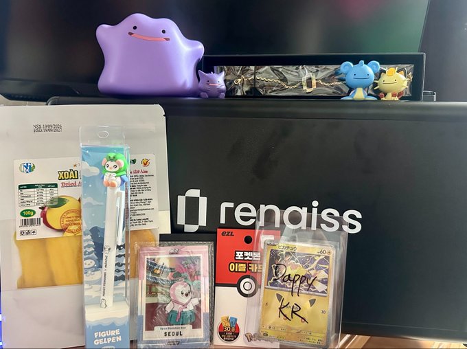 A collection of merchandise from a Renaiss (@renaissxyz) meetup is displayed on a desk, including a "Seoul" figure, a "Poppy KR" Pokémon card, "XOA Mango" snacks, a Korean chopsticks-and-fork set, a Pikachu figurine, and a purple Ditto plush resting on a Renaiss-branded laptop.