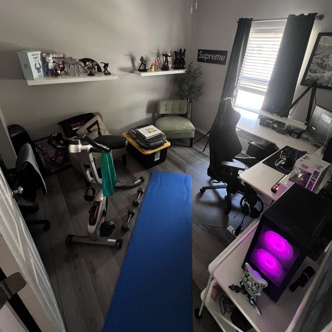 A view of a home office/gaming room featuring an exercise bike, a gaming chair at a desk with a PC, a blue yoga mat, floating shelves displaying figurines, and a 'Supreme' sign on the wall.
