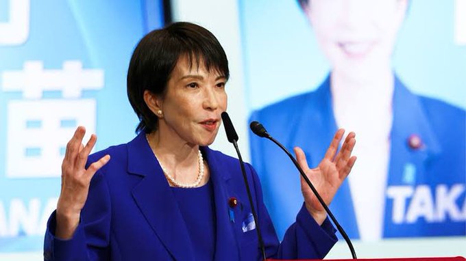Japanese politician Sanae Takaichi speaking at a podium during a press conference, with her name partially visible in Japanese on a blue background.