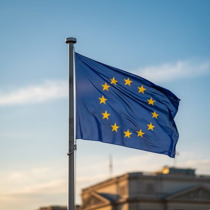 The European Union flag with its circle of yellow stars on a blue background, waving on a flagpole against a partly cloudy sky with a building visible below.