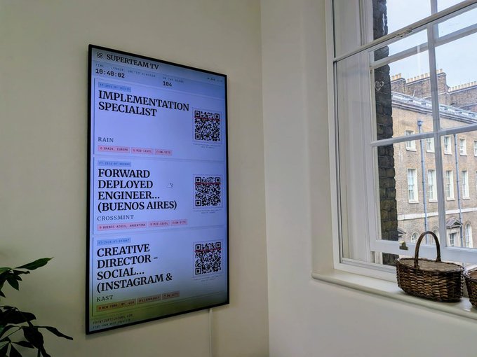 A digital display screen mounted on a white wall next to a window shows a 'SUPERTEAM TV' job board featuring listings like 'IMPLEMENTATION SPECIALIST' (RAIN), 'FORWARD DEPLOYED ENGINEER...' (CROSSMINT), and 'CREATIVE DIRECTOR - SOCIAL...' (KAST), each with a QR code.