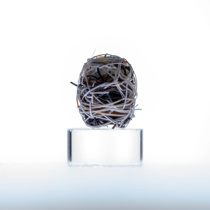 A spherical sculpture made of tangled, shredded paper strips in gray, black, and brown rests on a clear rectangular pedestal against a white background.