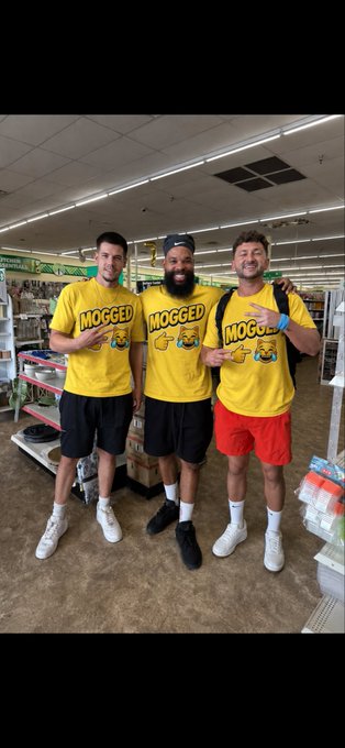 Three men pose together inside a store, all wearing matching yellow 'MOGGED' t-shirts and pointing to the logo.